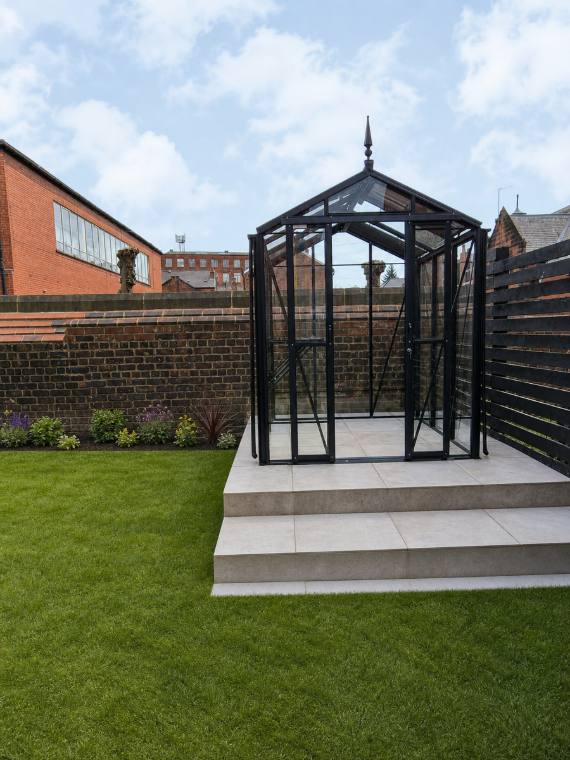 Black Victorian-style greenhouse with finial on a raised paved plinth in a walled urban garden